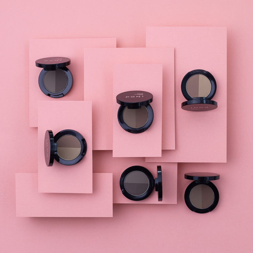 Several black round compacts of Brow Duo Powders by Poni Cosmetics are arranged on a pink background with overlapping rectangular pink sheets. Each compact is open, showcasing a duo of dark and light brown powders designed for thicker brows and defined arches. The arrangement is symmetrical and tidy.